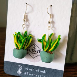 May include: Handmade earrings depicting miniature potted plants. Each earring features a green pot with a plant of yellow and green leaves. The earrings hang from silver-coloured hooks. The card behind the earrings has the text "FUENTE FINDS".