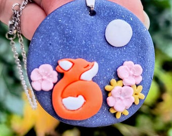 Könnte beinhalten: Eine runde Anhänger-Halskette mit einem skurrilen Design. Der Anhänger hat einen blauen Hintergrund mit einem weißen Mond, einem orangefarbenen Fuchs und rosa und gelben Blumen. Die Halskette hat eine silberne Kette.
