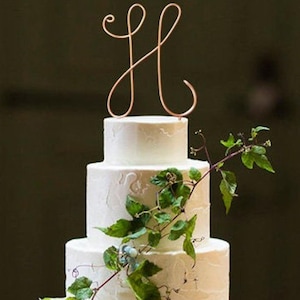 Peut inclure: Un gâteau de mariage blanc avec un topper en forme de lettre H cursive et des vignes vertes qui cascadent sur les côtés.