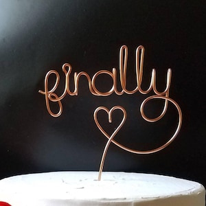 Könnte beinhalten: Ein goldfarbener Draht-Kuchentopper mit dem Wort "finally" und einem Herzen. Der Topper befindet sich auf einem weißen Kuchen.