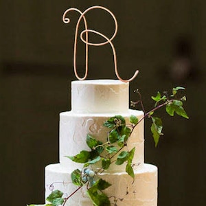 Peut inclure: Un gâteau de mariage blanc à trois étages décoré de vignes vertes et d'un topper en fil d'or en forme de lettre R.