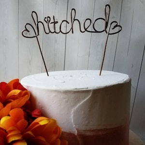 Könnte beinhalten: Ein weißer und rosa Kuchen mit einem Draht-Kuchentopper, auf dem "hitched" mit Herzen an beiden Enden steht.