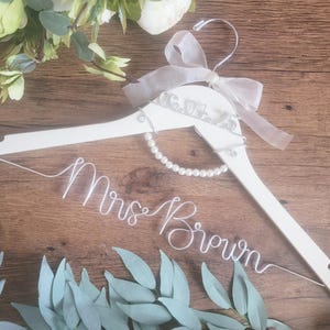 May include: A white wooden wedding dress hanger with a pearl necklace and a sheer ribbon bow. The hanger has the name "Mrs. Brown" in wire script. The date "06.09.23" is also on the hanger. Greenery and flowers are in the background.