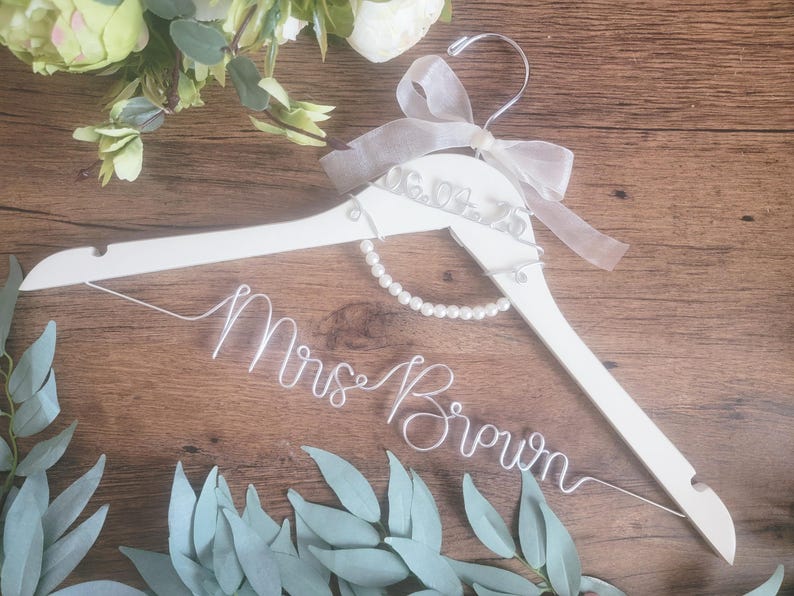 May include: A white wooden wedding dress hanger with a pearl necklace and a sheer ribbon bow. The hanger has the name "Mrs. Brown" in wire script. The date "06.09.23" is also on the hanger. Greenery and flowers are in the background.