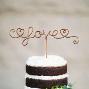 Könnte beinhalten: Ein Kuchentopper aus Kupferdraht mit dem Wort "love" und zwei Herzen auf jeder Seite. Der Topper befindet sich auf einer dreistöckigen Schokoladen-Torte mit weißem Frosting.