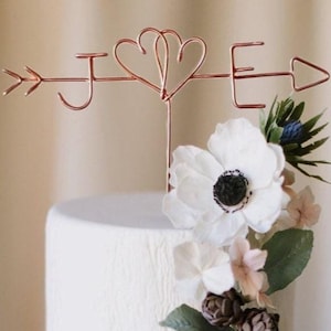 Könnte beinhalten: Eine weiße Hochzeitstorte mit einem Kuchentopper aus Kupferdraht, der die Buchstaben J und E mit einem Doppelherz-Design und einem Pfeil zeigt. Die Torte ist mit weißen und rosa Blumen verziert.