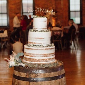 A rustic three tier naked wedding cake with a light gold wire cake topper with the script "The Marshalls".
