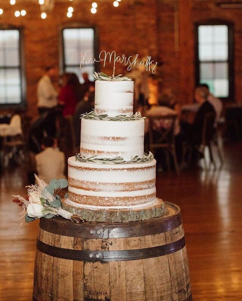 A rustic three tier naked wedding cake with a light gold wire cake topper with the script "The Marshalls".