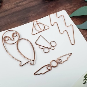 May include: A set of six gold-toned wire bookmarks featuring various shapes, including an owl, a lightning bolt, a triangle, a Hogwarts house crest, and a pair of wings.