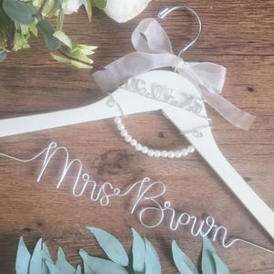May include: White wooden wedding dress hanger with the text "Mrs Brown" in wire. The hanger is decorated with a pearl strand, a sheer ribbon, and the date "06.04.23". Decorative flowers and greenery are in the background.