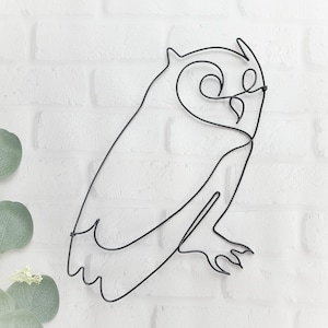 May include: A black wire sculpture of an owl with its wings outstretched. The owl is perched on a white brick wall.
