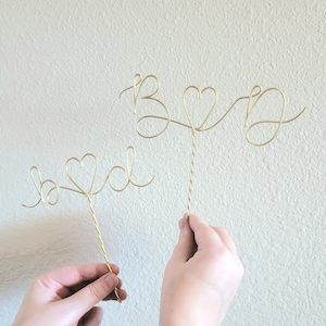 May include: Two gold wire cake toppers, one says "Bride" with a heart in the middle, the other says "Groom" with a heart in the middle.