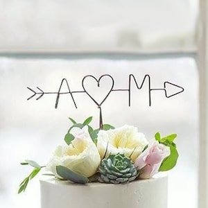 Könnte beinhalten: Ein Hochzeitstortenaufsatz aus Draht mit den Buchstaben A und M, die durch einen Pfeil mit einem Herz in der Mitte verbunden sind. Der Aufsatz befindet sich auf einer weißen Torte, die mit weißen und rosa Blumen und einer Sukkulente verziert ist.