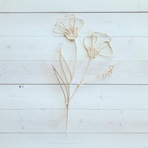 May include: Gold wire sculpture of two flowers with stems and leaves on a white wood background.
