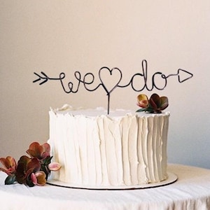 Könnte beinhalten: Weiße Hochzeitstorte mit einem schwarzen Draht-Tortenaufsatz, auf dem "we ♥ do" mit einem Pfeil auf jeder Seite steht.