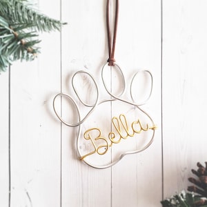 May include: A wire paw print ornament with the name "Bella" in gold script. The ornament hangs from a brown cord against a white wooden background, with pine accents.