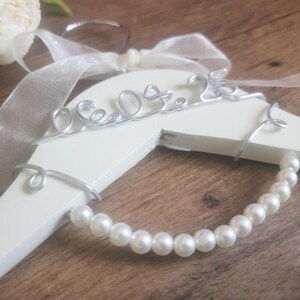 May include: A white wooden hanger with a silver wire design that reads "06.09.23". The hanger is adorned with a pearl strand and a white ribbon. The background is a wooden surface.
