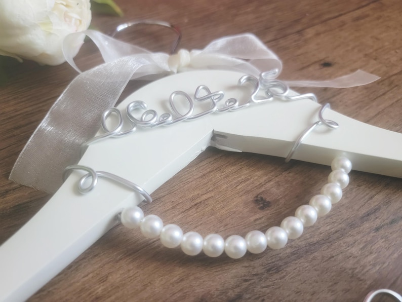 May include: A white wooden hanger with a silver wire design that reads "06.09.23". The hanger is adorned with a pearl strand and a white ribbon. The background is a wooden surface.
