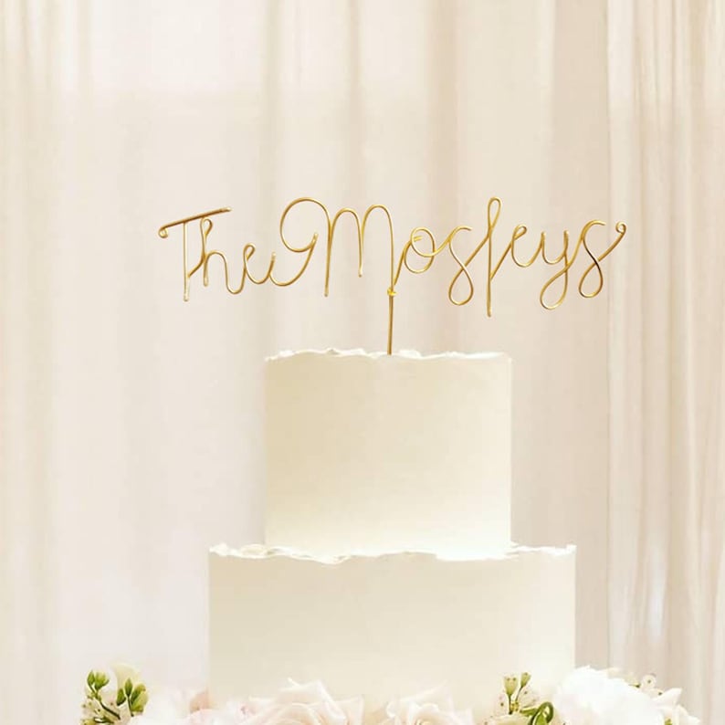Op de afbeelding: Een witte bruidstaart met twee lagen en een gouden taarttopper met de tekst "The Moseleys" in cursief schrift. De taart is versierd met witte glazuur en staat voor een witte gordijn.