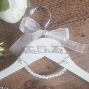 May include: A white wooden wedding dress hanger with a pearl bead strand and the date "06.04.25" in silver wire. A sheer white ribbon bow adorns the top, with a silver metal hook. A white rose and greenery are in the background.