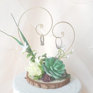 Può includere: Un portafedi a forma di cuore dorato con due fedi, decorato con una pianta grassa e fiori bianchi su una fetta di legno.