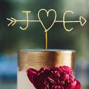 Könnte beinhalten: Goldener Kuchentopper aus Metall mit einem Herzen und den Buchstaben J und E auf beiden Seiten des Herzens. Die Buchstaben sind durch einen Pfeil mit einer Spitze an jedem Ende verbunden.