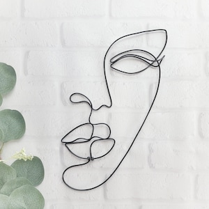 May include: Black wire sculpture of a face with a single eye and lips. The sculpture is mounted on a white brick wall.