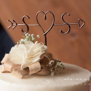 Könnte beinhalten: Eine weiße Hochzeitstorte mit einer rustikalen braunen Schleife aus Sackleinen und einer weißen Blume oben. Der Tortendecker ist ein Pfeil aus Metall mit den Buchstaben "S" und einem Herzen in der Mitte.