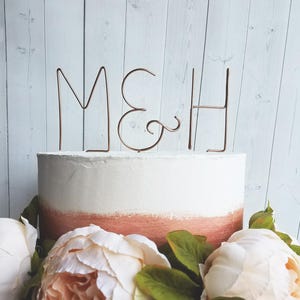 Könnte beinhalten: Eine weiße und pfirsichfarbene Ombré-Torte mit einem Kuchentopper aus Kupferdraht, auf dem "M&H" steht. Die Torte ist von weißen Pfingstrosen umgeben.