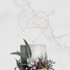 May include: A gold heart-shaped cake topper with the initials "P&P" and arrows on either side. The topper is on top of a gray cake decorated with greenery and flowers.
