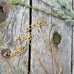 Può includere: Tre segnalibri per piante in filo dorato con le parole "Prezzemolo", "Salvia" e "Aglio" scritte su di essi. I segnalibri sono su una superficie di legno.