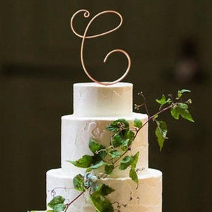Peut inclure: Un gâteau de mariage blanc à trois étages décoré d'une vigne feuillue et d'un topper en or en forme de lettre C cursive.