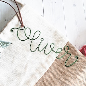 May include: A white and brown burlap stocking with a green wire name "Oliver" attached to the top.