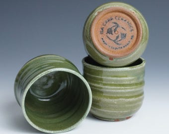 Könnte beinhalten: Vier handgefertigte grüne Keramikbecher. Die Becher haben ein strukturiertes, geriffeltes Design und eine dunkelgrüne Glasur. Auf dem Boden eines Bechers steht der Text "ISA CARR CERAMICS" und ein Fischlogo. Die Becher sind gestapelt.