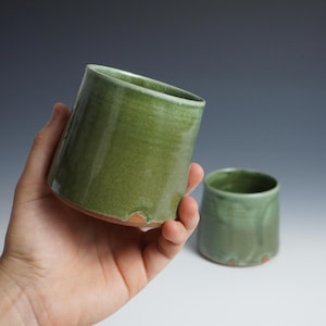 May include: Two handmade ceramic cups. One is held in a hand, the other sits on a white surface. Both are cylindrical with a green glaze and a reddish-brown base. The cups have a rustic, organic aesthetic.