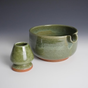May include: A green ceramic matcha bowl with a spout and a matching whisk holder. The bowl has a wide, shallow design with a reddish-brown base. The whisk holder is small and rounded, also with a reddish-brown base. Both items have a textured surface.