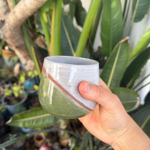 Könnte beinhalten: Ein handgefertigter Keramikbecher mit rundem Boden und geradem Rand. Die untere Hälfte ist ein gedämpftes Grün, das in ein graues Oberteil mit einem diagonalen braunen Streifen übergeht. Der Becher wird in einer Hand gehalten, mit einem Hintergrund aus grünen Pflanzen.