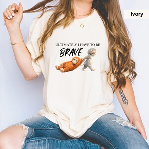 May include: Ivory-colored t-shirt featuring the text "ULTIMATELY I HAVE TO BE BRAVE" above an illustration of two cartoon monkeys. One monkey is lying down, while the other is pulling it along. The shirt is a relaxed fit.