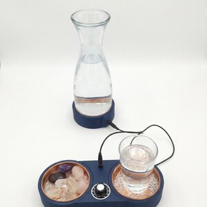 Könnte beinhalten: Eine klare Wasserkaraffe und ein Glas Wasser stehen auf einer blauen Basis mit kupferfarbenem Rand. Die Basis hat ein Fach mit Kristallen und einen Drehknopf. Eine zweite, kleinere Basis hält ein Glas Wasser.