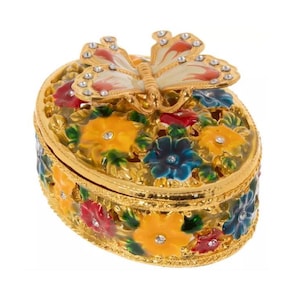 May include: An oval-shaped gold-tone metal trinket box featuring a raised butterfly on the lid. The exterior is decorated with colourful enamel flowers in yellow, red, and blue, accented with small clear crystals embedded in the floral centres.