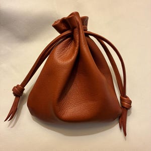 May include: A small, brown leather pouch with a drawstring closure. The pouch is gathered at the top and has a rounded bottom. The drawstring is tied in knots on each side. The leather has a textured appearance.