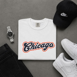 May include: White t-shirt with the word "Chicago" in a navy blue script with an orange outline. The shirt is paired with a black Nike baseball cap, white sneakers, a silver watch, and black jeans.