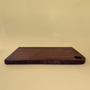 May include: A rectangular, dark brown wooden cutting board with a heart-shaped handle. The board has a smooth surface and rounded edges. The cutting board is approximately 12 inches long and 8 inches wide.