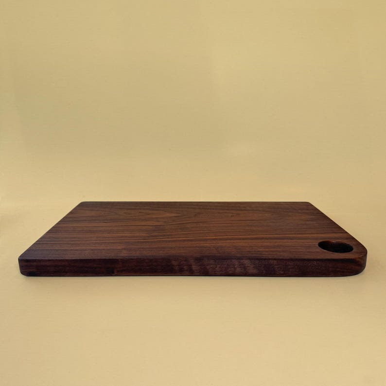 May include: A rectangular, dark brown wooden cutting board with a heart-shaped handle. The board has a smooth surface and rounded edges. The cutting board is approximately 12 inches long and 8 inches wide.