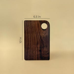 May include: A rectangular wooden cutting board with a dark brown finish. It measures 10 inches in height and 6.5 inches in width. A circular hole is cut out of the top right corner.