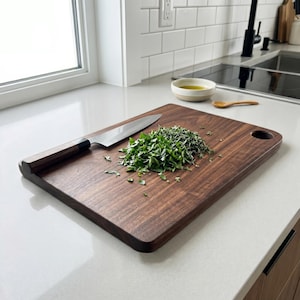May include: A rectangular, dark brown wooden cutting board with a rounded handle hole. A stainless steel chef's knife rests on the board next to a pile of chopped green herbs. The board is on a white countertop.