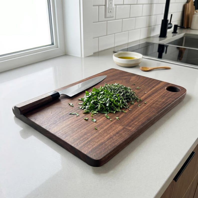 May include: A rectangular, dark brown wooden cutting board with a rounded handle hole. A stainless steel chef's knife rests on the board next to a pile of chopped green herbs. The board is on a white countertop.