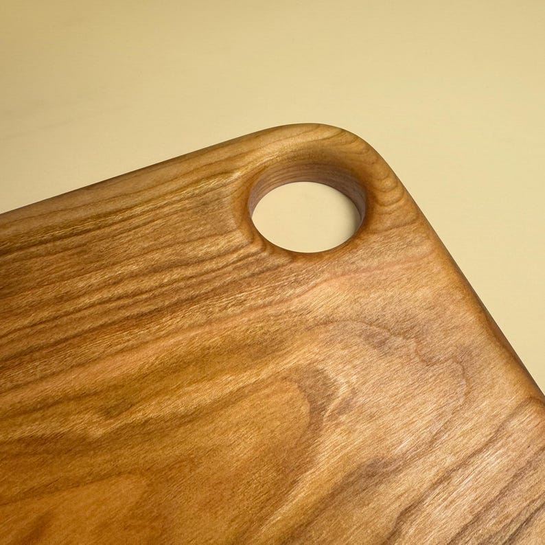 May include: Close-up of a wooden cutting board with a natural wood grain finish. The board has a rounded corner with a circular hole for hanging or gripping. The wood has warm brown tones.