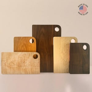 May include: A collection of wooden cutting boards in various sizes and wood tones, including light maple, walnut, and cherry. Each board has a rounded corner with a circular handle. The image includes a "Made in USA" sticker.