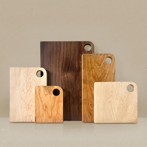 May include: A collection of wooden cutting boards in various sizes and wood grains. The boards range in color from light maple to dark walnut, each with a rounded corner and a circular handle hole.